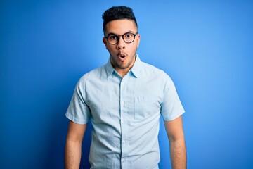Young handsome man wearing casual summer shirt and glasses over isolated blue background afraid and shocked with surprise expression, fear and excited face.