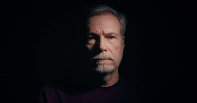 A mature bearded man emerging from the darkness to a sole light source that illuminates his somber face.
