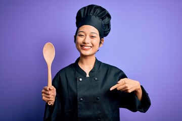 Fototapeta premium Young beautiful chinese chef woman wearing cooker uniform and hat holding wooden spoon with surprise face pointing finger to himself