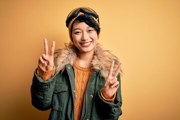 Beautiful asian skier girl wearing snow sportswear using ski goggles over yellow background smiling looking to the camera showing fingers doing victory sign. Number two.