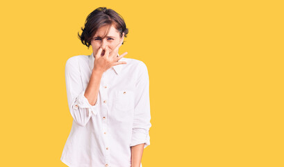 Beautiful young woman with short hair wearing elegant white shirt smelling something stinky and disgusting, intolerable smell, holding breath with fingers on nose. bad smell