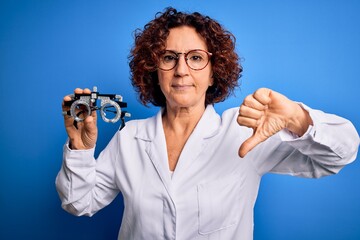 Middle age optical woman wearing coat holding optometry glasses over blue background with angry face, negative sign showing dislike with thumbs down, rejection concept