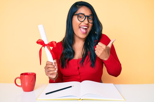 Beautiful latin young woman with long hair holding graduate degree diploma pointing thumb up to the side smiling happy with open mouth