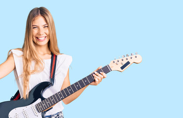 Fototapeta premium Beautiful caucasian woman with blonde hair playing electric guitar screaming proud, celebrating victory and success very excited with raised arms