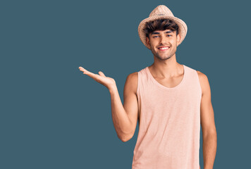Young hispanic man wearing summer hat smiling cheerful presenting and pointing with palm of hand...