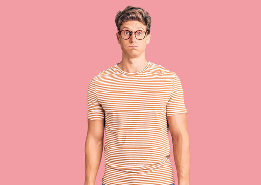 Young handsome man wearing casual clothes and glasses puffing cheeks with funny face. mouth inflated with air, crazy expression.