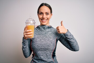 Young fitness woman wearing sport workout clothes drinking fresh orange juice happy with big smile doing ok sign, thumb up with fingers, excellent sign