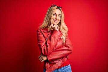 Fototapeta premium Young beautiful blonde woman wearing casual jacket standing over isolated red background looking confident at the camera with smile with crossed arms and hand raised on chin. Thinking positive.