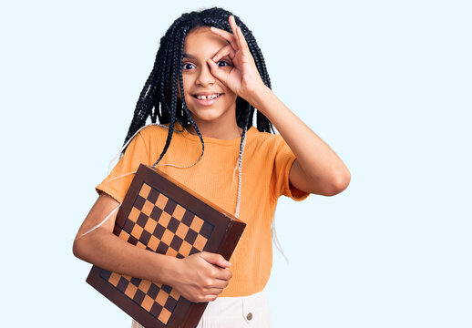 Cute african american girl holding chess smiling happy doing ok sign with hand on eye looking through fingers