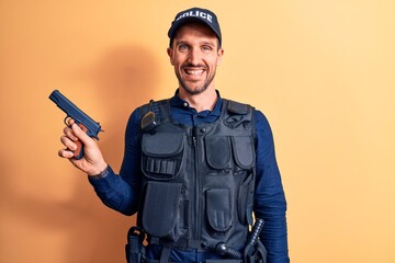 Handsome policeman wearing uniform and bulletprof holding gun over yellow background looking positive and happy standing and smiling with a confident smile showing teeth