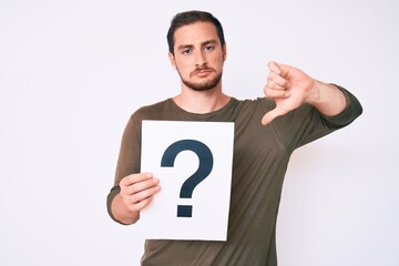 Young handsome man holding question mark with angry face, negative sign showing dislike with thumbs...