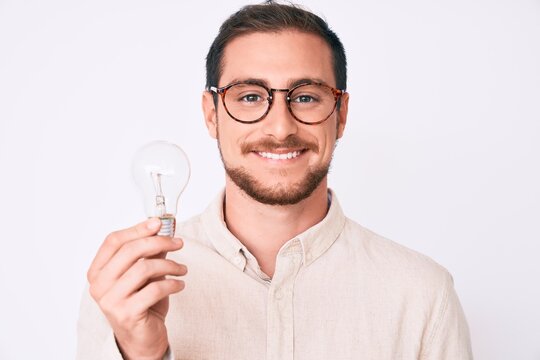 Young handsome man holding lightbulb for inspiration and idea looking positive and happy standing and smiling with a confident smile showing teeth