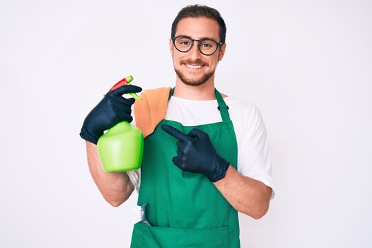 Young handsome man wearing apron holding sprayer smiling happy pointing with hand and finger