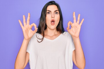 Young beautiful brunette woman wearing casual white t-shirt over purple background looking surprised and shocked doing ok approval symbol with fingers. Crazy expression