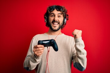 Fototapeta na wymiar Young gamer man with curly hair and beard playing video game using joystick and headphones screaming proud and celebrating victory and success very excited, cheering emotion