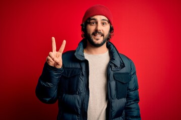 Young handsome man with beard wearing casual coat and wool cap over red background smiling with happy face winking at the camera doing victory sign. Number two.