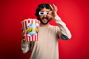 Young handsome man with beard watching movie holding popcorns over red background with happy face smiling doing ok sign with hand on eye looking through fingers