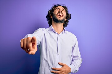 Young handsome business man with beard wearing shirt standing over purple background laughing at you, pointing finger to the camera with hand over body, shame expression