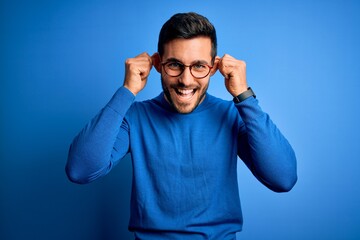 Naklejka premium Young handsome man with beard wearing casual sweater and glasses over blue background Smiling pulling ears with fingers, funny gesture. Audition problem