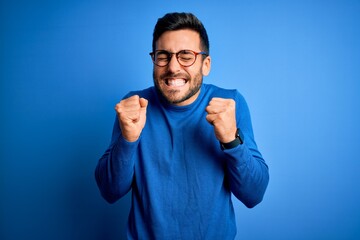 Young handsome man with beard wearing casual sweater and glasses over blue background excited for...