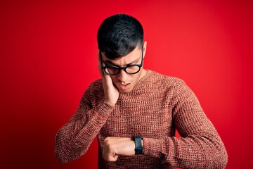 Young handsome hispanic man wearing nerd glasses over red background Looking at the watch time...