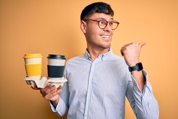 Young scholarship holder business man holding take away coffee over isolated yellow background pointing and showing with thumb up to the side with happy face smiling