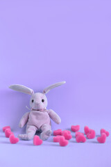 Pink plush hare sits on violet background among soft pink hearts, vertical orientation. Valentine's day card