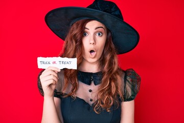 Young beautiful woman wearing witch halloween costume holding paper with trick or treat message scared and amazed with open mouth for surprise, disbelief face