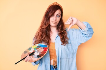 Young beautiful woman holding paintbrush and palette with angry face, negative sign showing dislike with thumbs down, rejection concept