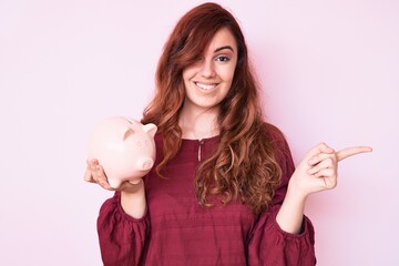 Young beautiful woman holding piggy bank smiling happy pointing with hand and finger to the side
