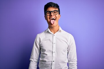 Obraz premium Young handsome business man wearing shirt and glasses over isolated purple background sticking tongue out happy with funny expression. Emotion concept.