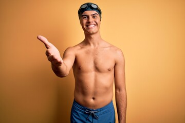 Young handsome man shirtless wearing swimsuit and swim cap over isolated yellow background smiling friendly offering handshake as greeting and welcoming. Successful business.