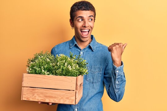 Young african amercian man holding wooden pot with plant pointing thumb up to the side smiling happy with open mouth