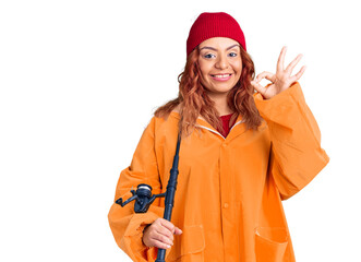 Young latin woman wearing fisher raicoat holding rod doing ok sign with fingers, smiling friendly gesturing excellent symbol