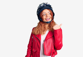 Young latin woman wearing leather jacket holding motorcycle helmet smiling with happy face looking and pointing to the side with thumb up.