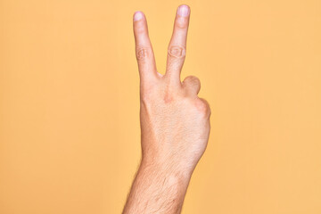 Hand of caucasian young man showing fingers over isolated yellow background counting number 2 showing two fingers, gesturing victory and winner symbol
