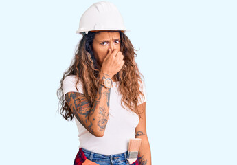 Young hispanic woman with tattoo wearing hardhat and builder clothes smelling something stinky and disgusting, intolerable smell, holding breath with fingers on nose. bad smell