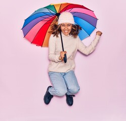 Young beautiful latin woman holding umbrella smiling happy. Jumping with smile on face over isolated pink background