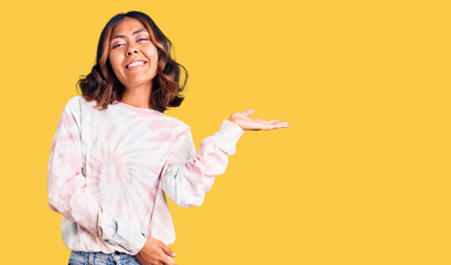 Young beautiful mixed race woman wearing casual tie dye sweatshirt smiling cheerful presenting and pointing with palm of hand looking at the camera.