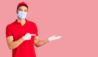 Young handsome hispanic man wearing delivery uniform and medical mask amazed and smiling to the camera while presenting with hand and pointing with finger.