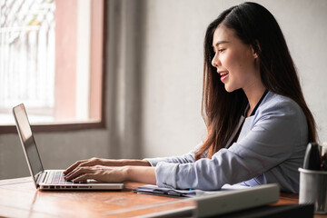 Young happy Asian businesswoman in blue shirt working from home and use a computer laptop and thinking idea for her business