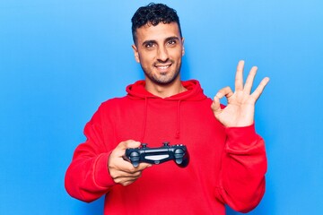 Young hispanic man playing video game holding controller doing ok sign with fingers, smiling friendly gesturing excellent symbol