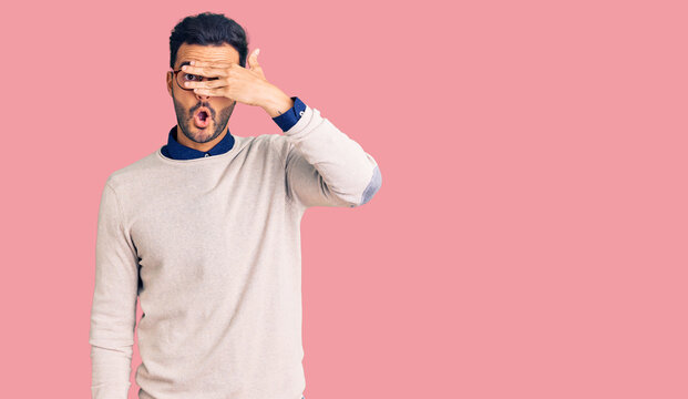 Young handsome hispanic man wearing elegant clothes and glasses peeking in shock covering face and eyes with hand, looking through fingers with embarrassed expression.