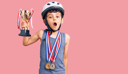 Little cute boy kid wearing bike helmet and winner medals holding winner trophy scared and amazed with open mouth for surprise, disbelief face