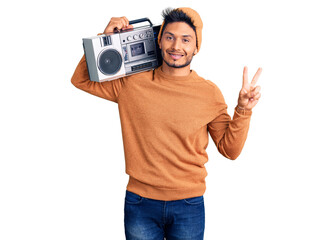 Handsome latin american young man holding boombox, listening to music smiling looking to the camera showing fingers doing victory sign. number two.