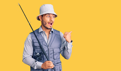 Handsome latin american young man wearing fisherman equipment pointing thumb up to the side smiling happy with open mouth