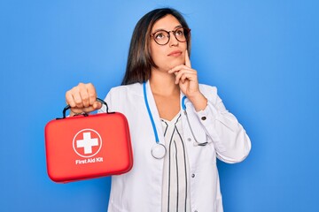 Young doctor woman holding medical first aid kit red box for emergency over blue background serious...