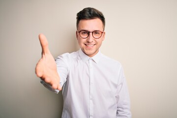 Young handsome business mas wearing glasses and elegant shirt over isolated background smiling friendly offering handshake as greeting and welcoming. Successful business.