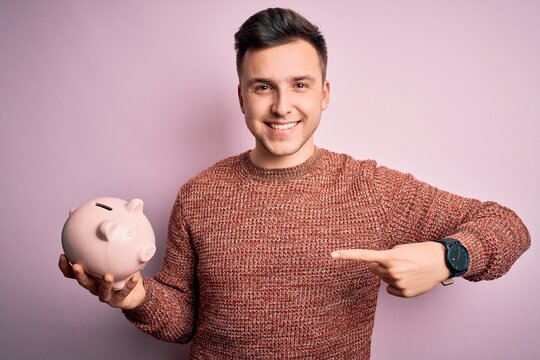 Young Handsome Caucasian Man Holding Piggy Bank For Savings Over Pink Background With Surprise Face Pointing Finger To Himself