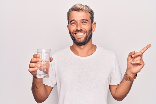 Handsome blond man with beard drinking glass of water to refreshment over white background smiling happy pointing with hand and finger to the side